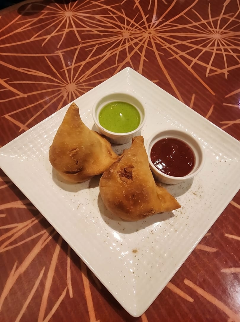 Two golden-brown samosas on a white rectangular plate with green and red chutneys.