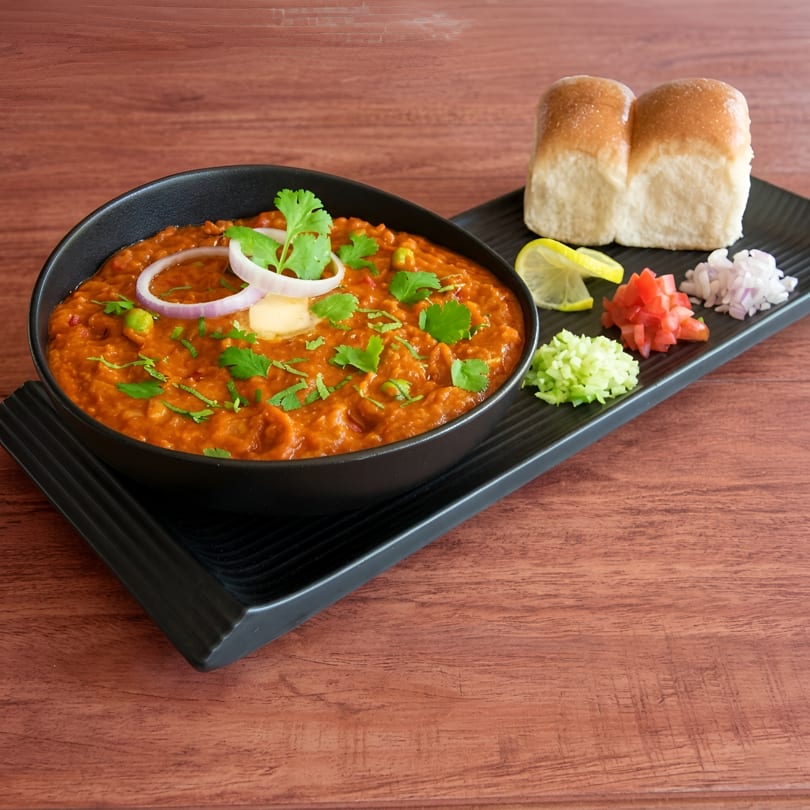 A black bowl of pav bhaji with butter, onions, and cilantro, served with pav and chopped vegetables.