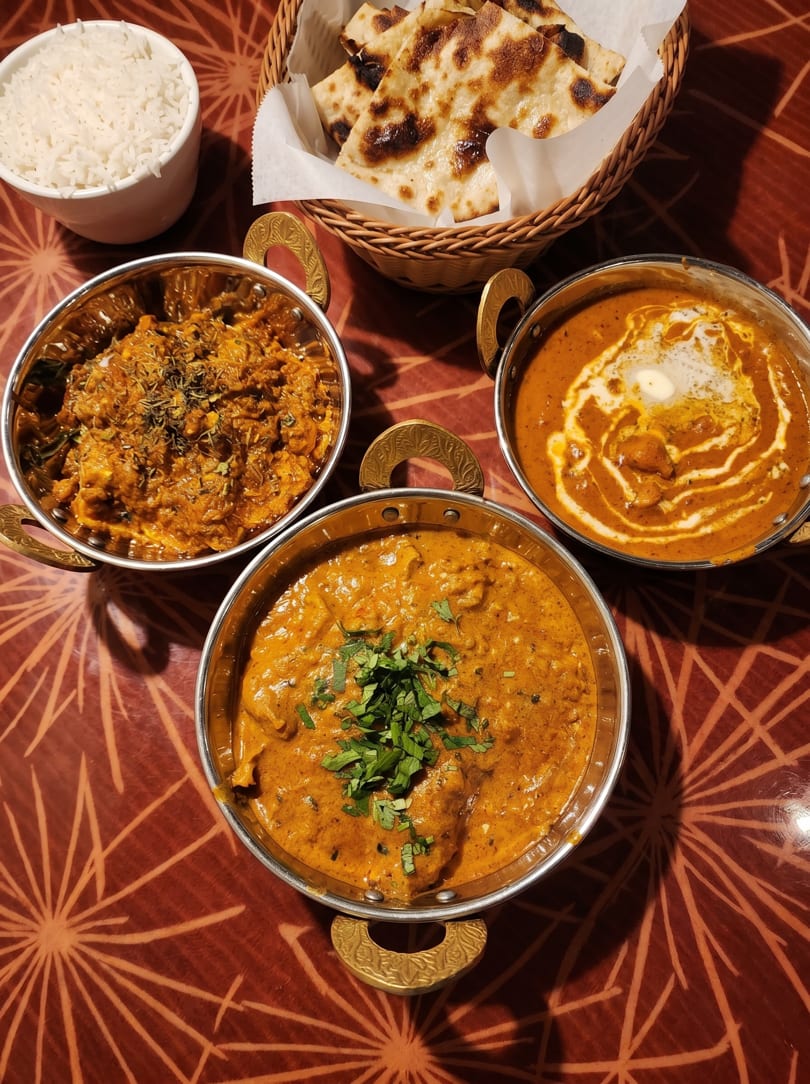 Three bowls of Indian curry, a basket of naan bread, and a cup of rice on a patterned table.