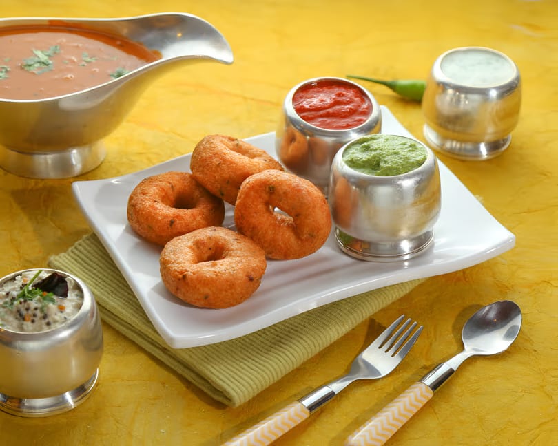 Golden-brown vada donuts on a white plate with various chutneys and sambar in silver bowls.