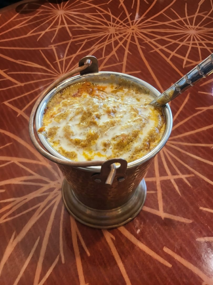 A copper bucket filled with creamy Indian curry, drizzled with white sauce, on a patterned red table.