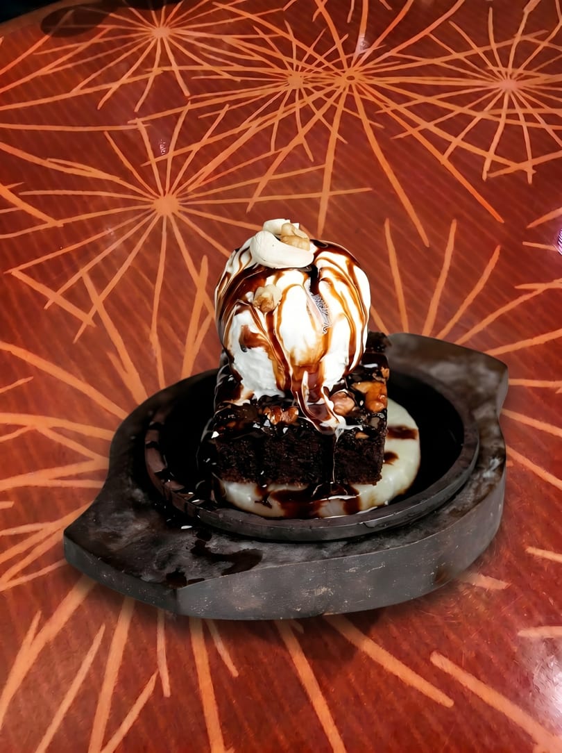 A sizzling brownie with vanilla ice cream, chocolate sauce, and nuts on a dark plate.