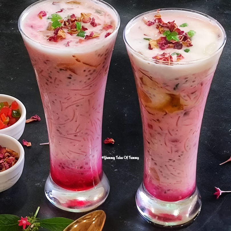 Two tall glasses of pink falooda, topped with cream, rose petals, and green garnish.