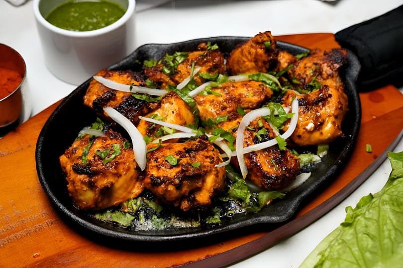 Sizzling chicken tikka with onions and cilantro on a black skillet, with green and red sauces.