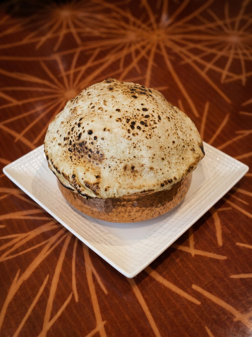 A golden pot of biryani, sealed with a charred flatbread, sits on a white plate.