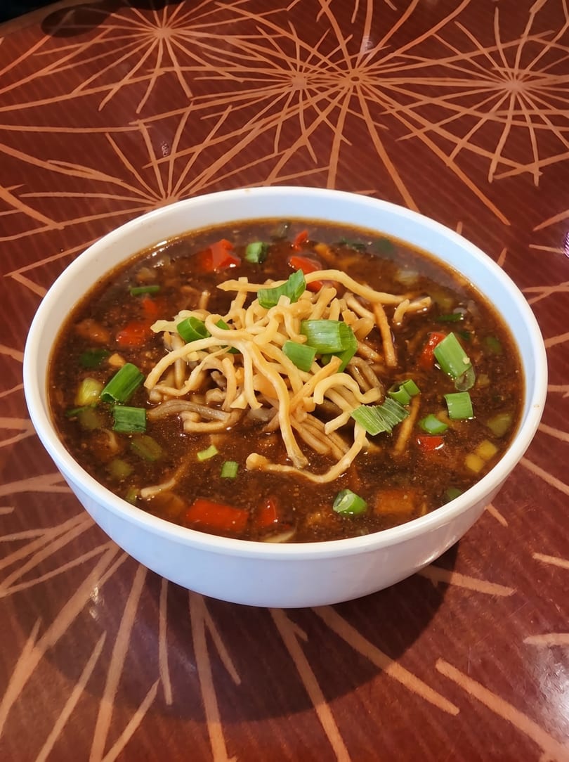 A white bowl of dark soup with noodles, red peppers, and green onions on a patterned table.