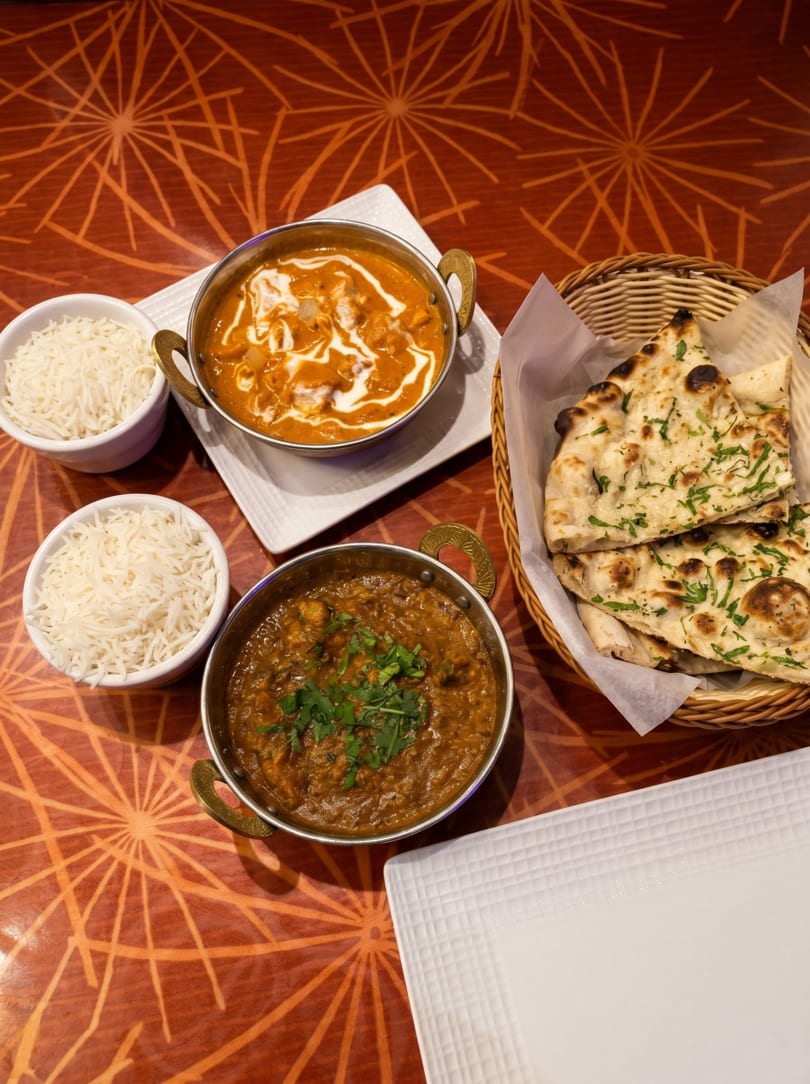 Indian meal with two curries, basmati rice, and naan bread on a patterned orange table.